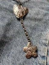 Collar metálico plateado con dije de corazón y de flor. La cadena está formada por dos modelos distintos: una con caritas sonrientes y otra de eslabones gruesos.