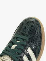Zapatillas Adidas Samba OG con exterior de piel con estampado de piel de serpiente en tonos verde oscuro y negro. Cuentan con las 3 Franjas dentadas en color blanco crema, cordones verde oscuro, revestimientos en color crema claro y puntera en T reforzada. La suela es de caucho color caramelo.