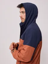 Campera rompevientos con cierre frontal, confeccionada en nylon con diseño de bloques de color en tonos verde azulado y gris claro. Cuenta con capucha, puños elásticos y cordón de ajuste en el ruedo.