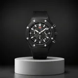 Reloj de pulsera analógico con cronógrafo, caja redonda de metal negro y correa de caucho negra. Presenta esfera texturizada con marcadores luminosos y detalles en rojo.