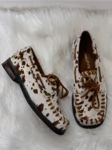 Zapato tipo mocasín de cuero con pelo estampado con diseño de manchas de vaca en tonos marrón y blanco. Presenta cordones frontales, costuras visibles en el borde y suela plana.