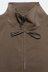 Chaleco fluido color marrón con cuello subido, sin mangas. Presenta cierre frontal de cremallera metálica y detalles de lazadas ajustables en el cuello y la cintura.