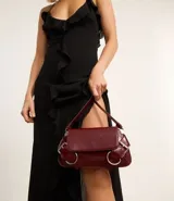 Cartera tipo baguette de tamaño pequeño, confeccionada en poliuretano color negro. Presenta una solapa frontal con detalles de hebillas metálicas decorativas en los laterales y un asa de hombro ajustable.