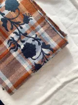Bufanda de gran tamaño con diseño de cuadros en tonos naranja, beige y azul, decorada con un bordado floral en color azul oscuro y terminaciones con flecos.