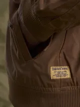 Campera tipo bomber color marrón, confeccionada en tela encerada, con cierre frontal, cuello tipo mao y puños y ruedo acanalados.
