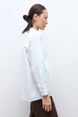 Camisa blanca de manga larga con cuello clásico y botones frontales. Presenta dos bolsillos asimétricos en el pecho, uno con solapa y otro sin.