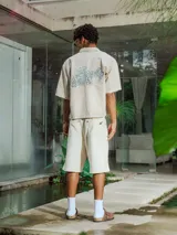 Camisa de manga corta color crudo, confeccionada en gabardina, con cuello camisero y botones frontales. Presenta un bolsillo de parche en el pecho y bordado en la espalda.