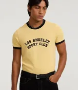 Remera de manga corta estilo ringer, color amarillo claro con ribetes negros en cuello y mangas. Presenta un estampado central en negro con el texto "Los Angeles Sport Club".