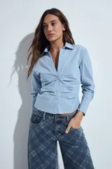 Camisa blanca de manga larga con cuello clásico y cierre frontal de botones. Presenta un diseño entallado con frunces decorativos en la parte central delantera.