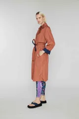 Trench largo cruzado color ladrillo, sin forro, con cinturón reversible azul marino y detalles de color de contraste ocultos. Tiene botones en el frente, bolsillos de vivo oblicuos y capucha oculta.