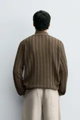 Polo de punto color marrón oscuro, de calce regular, con cuello solapa y abertura frontal en V. Presenta tejido con trencitas verticales.