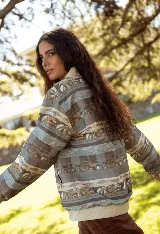 Campera de jean con cierre frontal y cuello tipo bomber, presenta un estampado horizontal con motivos paisajísticos y geométricos en tonos azules, marrones y beige. Posee puños de tela elástica en color blanco.