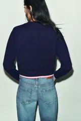 Chaqueta de punto de cuello redondo y manga larga, con cierre frontal de botones. Presenta dos bolsillos delanteros de plastrón y detalles de ribetes en color rosa a contraste en los puños, el ruedo y los bolsillos.