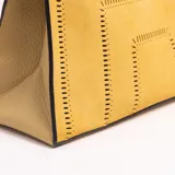 Cartera tipo tote de eco-cuero gamuzado color mostaza, con diseño estructurado de base trapezoidal y detalles calados geométricos. Cuenta con asa superior corta.