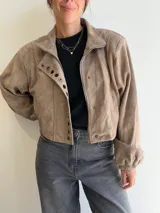 Chaqueta corta de efecto gamuza con cierre frontal y botones. Presenta mangas tipo globo con puños elásticos y detalle de aros metálicos decorativos en la solapa. Cuenta con ajuste lateral en la cintura y forro interior.