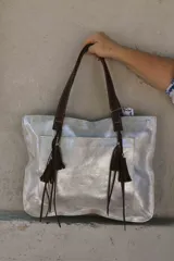 Bolso shopper plateado con textura craquelada, doble asa de cuero marrón con detalle de borlas y bolsillo externo.