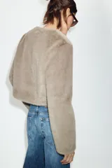 Chaqueta corta de cuello redondo y manga larga, confeccionada en tejido sintético con acabado efecto pelo. Presenta cierre frontal con botones a tono.