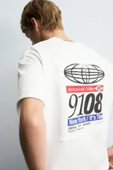 Camiseta blanca de corte boxy fit, cuello redondo y manga corta. Presenta un parche bordado con las letras "NY" y un diseño abstracto en negro en el pecho.