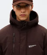 Campera tipo parka color marrón con capucha estructurada, cierre frontal y bolsillo vertical con cierre en el pecho. Presenta detalles de estampado utilitario en el frente y recortes en el cuerpo.