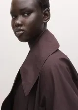 Chaqueta larga tipo gabardina color marrón oscuro, de diseño recto, con solapas amplias y cinturón ajustable del mismo tejido.
