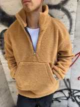 Buzo de corderito color beige, con cuello alto y cierre frontal parcial. Presenta un bolsillo tipo canguro frontal y diseño de corte holgado.