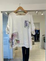 Blusa blanca de lino con detalles estampados florales en el bolsillo del pecho y en el bajo lateral. Tiene cuello redondo y mangas cortas con presilla y botón para ajustar.