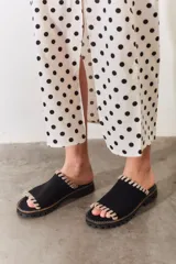 Chancla tipo mule o slider de color negro, con tira ancha sobre el empeine. El borde de la tira está decorado con un pespunte grueso de hilo color beige o crudo, creando un efecto trenzado o de festón.