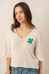 Sweater de tejido de punto fino color beige, con escote en V, mangas cortas holgadas y detalle de trébol verde bordado en el pecho.