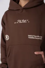 Buzo canguro color marrón con capucha, confeccionado en frisa de algodón. Presenta un diseño gráfico estampado en el frente con texto y logo circular, bolsillo tipo canguro y puños acanalados.