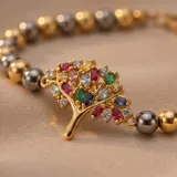 Pulsera de acero con baño de oro, compuesta por esferas doradas y oscuras, con un dije central en forma de árbol de la vida decorado con cristales multicolores.
