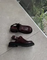 Zueco de cuero vacuno color bordó, con diseño de punta cuadrada y tira trasera ajustable con hebilla metálica. Presenta detalles de herrajes en tono oro viejo y una plataforma de goma negra con suela dentada.