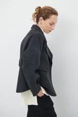 Chaqueta de paño color gris oscuro, con solapas amplias y cierre frontal mediante un alamar único. Presenta bolsillos laterales y mangas largas con puños rectos.