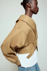 Chaqueta corta de Zara confeccionada en mezcla de algodón, con cuello solapa, manga larga con hombro caído y puños elásticos. Presenta un diseño cruzado con cierre de hebilla metálica lateral y forro interior a tono.