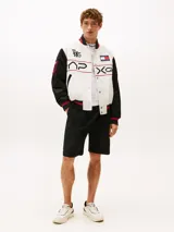 Campera estilo universitario de Tommy Hilfiger, colección APXGP, color blanco con mangas negras y detalles en rojo. Confeccionada en satén aterciopelado, con cuello estilo béisbol, cierre a presión, bolsillos laterales ribeteados, cuello, puños y bajo de punto canalé con rayas, logo de la colección APXGP bordado en la parte delantera y parches con el logo de la colección APXGP en mangas.