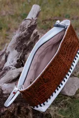 Sobre tipo baguette de cuero con textura croco en color marrón. Presenta un diseño bicolor con un lateral en cuero blanco unido mediante un trenzado artesanal en color negro. Incluye una etiqueta de cuero con el logo de la marca y un lazo desmontable.