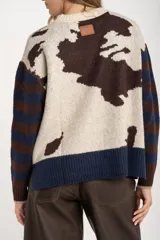 Sweater de punto con diseño combinado, presentando un estampado de manchas de vaca en el cuerpo y mangas con rayas horizontales en tonos azul y marrón. Posee cuello redondo y puños acanalados.