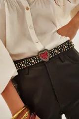 Cinturón elástico negro con hebilla en forma de corazón recubierta de strass rojos. La correa presenta aplicaciones de tachas metálicas y cristales en forma de estrellas y círculos.