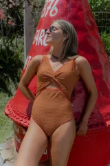 Malla enteriza color marrón con textura de relieve, escote en V con detalle de nudo frontal y aberturas laterales. Presenta bordes en color rojo contrastante.