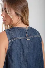 Blusa sin mangas de denim lavado oscuro, con cuello redondo y costuras verticales contrastantes en color camel. Presenta un dobladillo deshilachado y un pequeño parche rectangular con diseño gráfico multicolor en la parte inferior frontal.