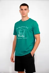 Remera de manga corta color verde esmeralda con cuello redondo. Estampado frontal en blanco que incluye un dibujo de una casa rodante (campervan) bajo palmeras y el texto "The Road Never Ends" y "Campervan Forever".