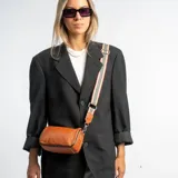 Cartera bandolera compacta de eco cuero color suela, con forma cilíndrica y correa ancha regulable de diseño rayado en tonos beige, negro y rojo.
