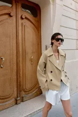 Trench corto de color beige, con cuello de solapa, manga larga con hombreras y trabillas con botones. Cuenta con cierre frontal cruzado con botones oscuros y cinturón ajustable del mismo tejido.