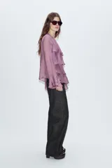 Blusa de cuello redondo y manga larga, color marrón oscuro. Presenta volantes en el frente y en los puños, y un bajo asimétrico. Cierre en la espalda con abertura y botón forrado.