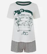 Pijama corto de viscolycra, compuesto por una blusa de manga corta color crema con cuello redondo y ribete verde, y un estampado frontal de un perro durmiendo en un sofá con la frase "Dog Dreams". El short es de corte recto, con cintura elástica y estampado de rayas rosas y verdes.