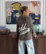 Sweater de textura suave y afelpada en color gris oscuro, con diseño de cuello redondo y mangas largas. Presenta un estampado gráfico en color blanco en la espalda con la inscripción 'odd studios'.