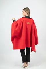 Capa de paño color rojo con corte amplio y caída natural. Presenta detalles de dijes metálicos en baño de oro distribuidos en la espalda y una etiqueta de cuero con el logo de la marca en la parte superior.