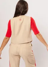 Chaleco beige con escote en V, sin mangas y cierre frontal con botones. Presenta pespuntes en color rojo y logo bordado en la espalda.