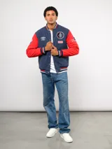 Campera bomber estilo universitario de Umbro, con cuerpo en azul marino y mangas rojas. Presenta cierre frontal con botones a presión, cuello, puños y ruedo con tejido acanalado tricolor, y diversos parches bordados alusivos al Club Nacional de Football.