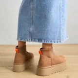 Botas cortas tipo plataforma UGG modelo Mini Dipper, confeccionadas en gamuza color tostado con panel superior de neopreno elástico y tirador naranja en el talón.