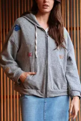 Campera con capucha y cierre frontal, confeccionada en algodón gris jaspeado. Presenta detalles decorativos de figuras como corazones, estrellas y coronas con acabado brillante en diversos colores. Cuenta con bolsillos laterales y cordones ajustables en la capucha.